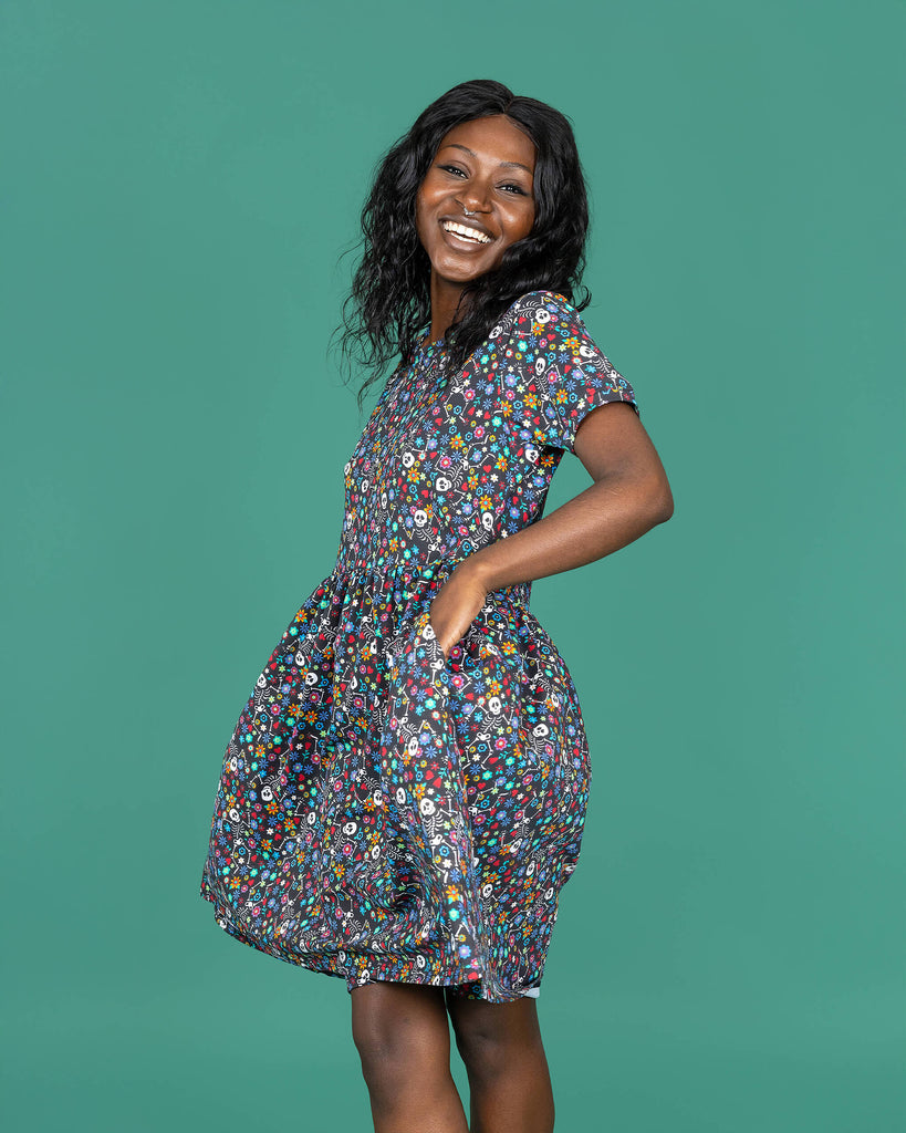 Close up of the Boogie Bones Oversized Smock Dress worn by a femme alternative model with mid length black hair on a teal studio background. She is facing forward swishing the dress with both hands in the dress pockets laughing to camera. The black base dress features dancing smiling skeletons with multicoloured flowers in various sizes and types all over.