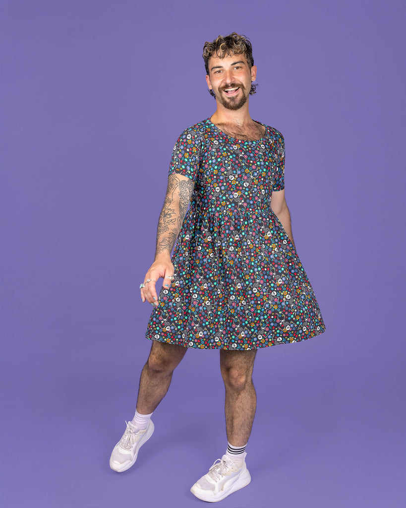 The Boogie Bones Oversized Smock Dress on a masc non binary model with a short curly brown mullet and white trainers on a purple studio background. They are facing forward laughing swishing the dress with their arms out to their side. The black base dress features dancing smiling skeletons with multicoloured flowers in various sizes and types all over.