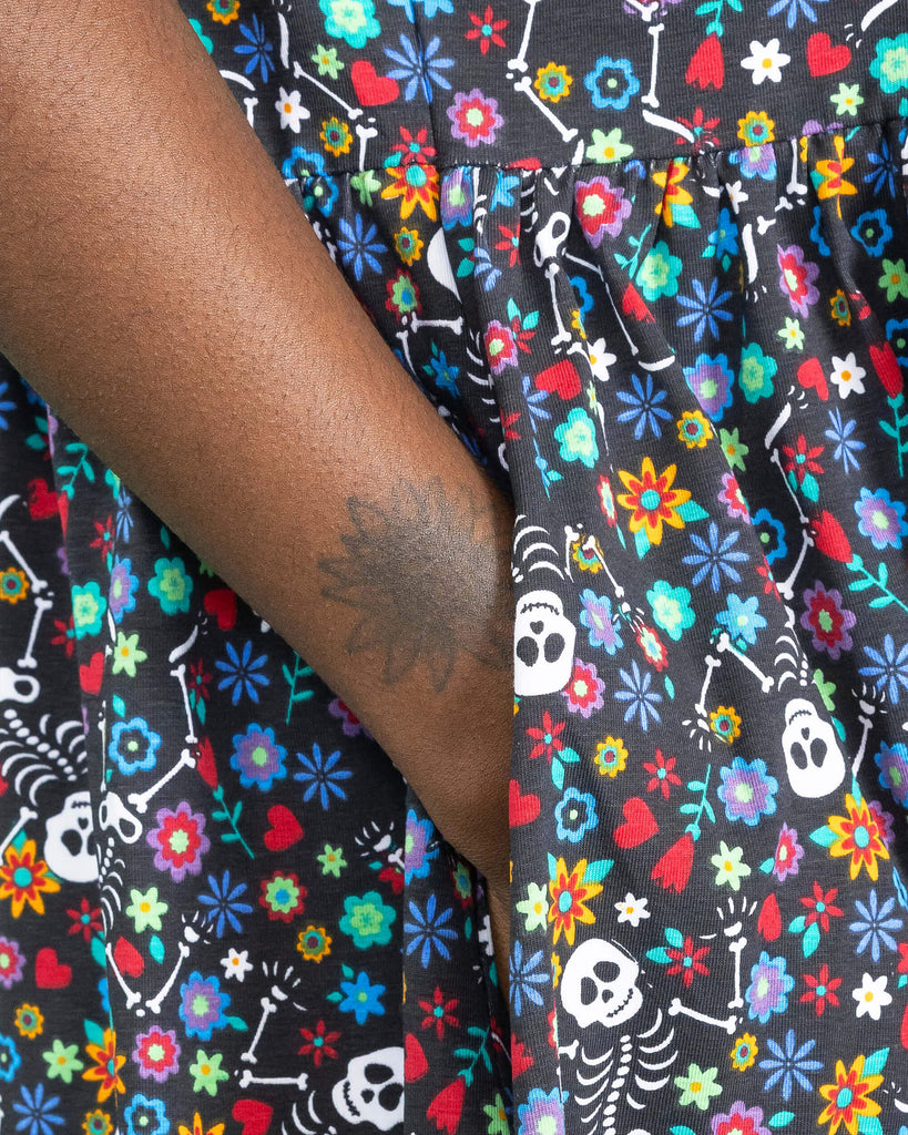 Close up of the Boogie Bones Oversized Smock Dress worn by a femme alternative model. She is facing right with her hand resting in the side dress pocket. The black base dress features dancing smiling skeletons with multicoloured flowers in various sizes and types all over.