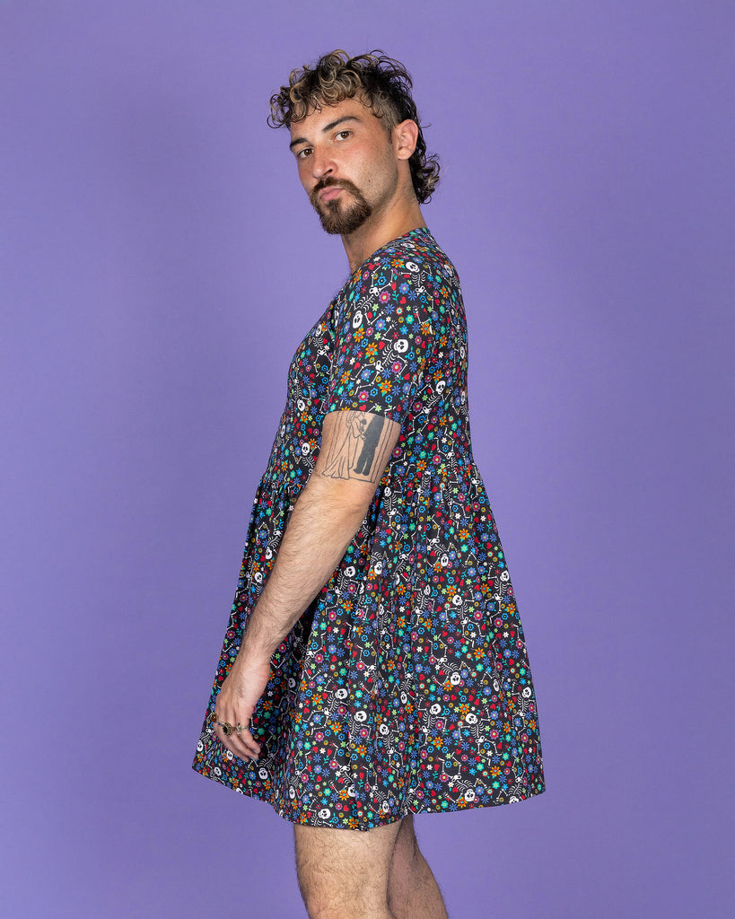 The Boogie Bones Oversized Smock Dress on a masc non binary model with a short curly brown mullet and white trainers on a purple studio background. They are facing left looking and pouting to camera with their hands resting by their sides. The black base dress features dancing smiling skeletons with multicoloured flowers in various sizes and types all over.