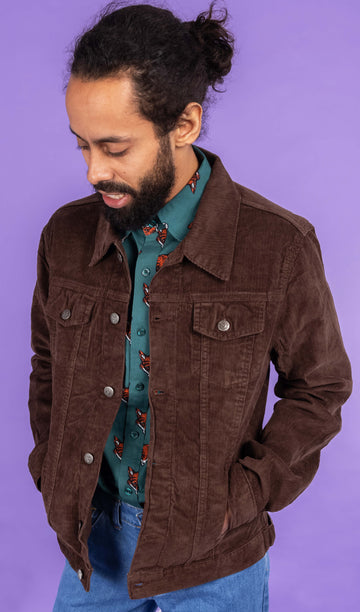 Richard, a hispanic male model with dark hair in a bun and a beard, is stood in a photography studio in Hove in front of a lilac backdrop wearing Retro Vintage Cocoa Brown Corduroy Unisex Western Jacket with Preppy Foxy Short Sleeve Shirt underneath and blue jeans. Richard is posing to one side and is looking down with his hands in the pockets of the jacket. The photo is cropped at the thigh.