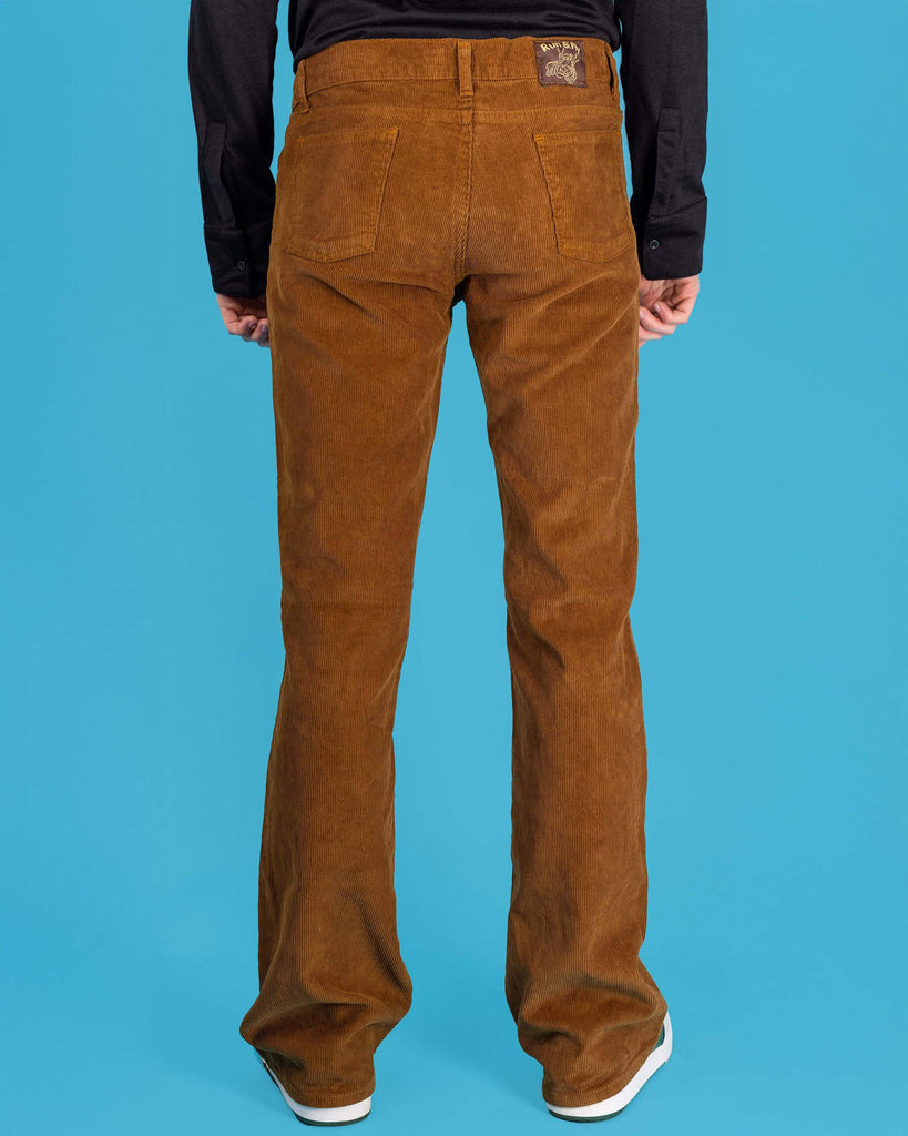 Jack is stood in front of a blue studio background wearing the tobacco cord bootcut flares with a long sleeve black tshirt and white trainers. They are facing away from the camera with both arms resting by their sides. Photo is cropped from the waist down.