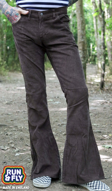 Model is stood in a forest area wearing the brown corduroy bell bottom super flares with a navy and white stripe t-shirt and checkered van shoes. They have one hand in their pocket with one hand behind their back, they are stood facing the camera with one leg slightly out to the side to show off the flares. Photo is cropped from the waist down.