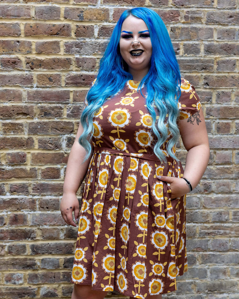 Georgina a blue long haired white plus sized model wearing a brown retro tea dress with floral print against a brick wall