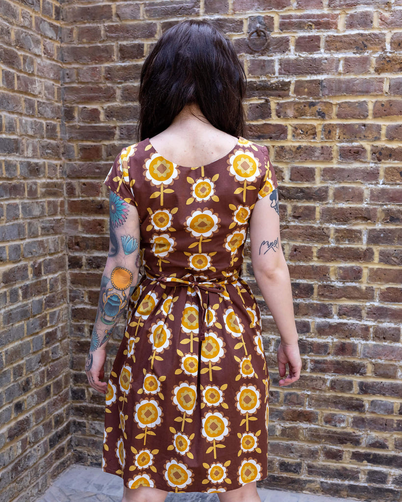 Shot of the back of Florence a brown long haired white model with tattoos is wearing a brown retro tea dress with floral print against a brick wall