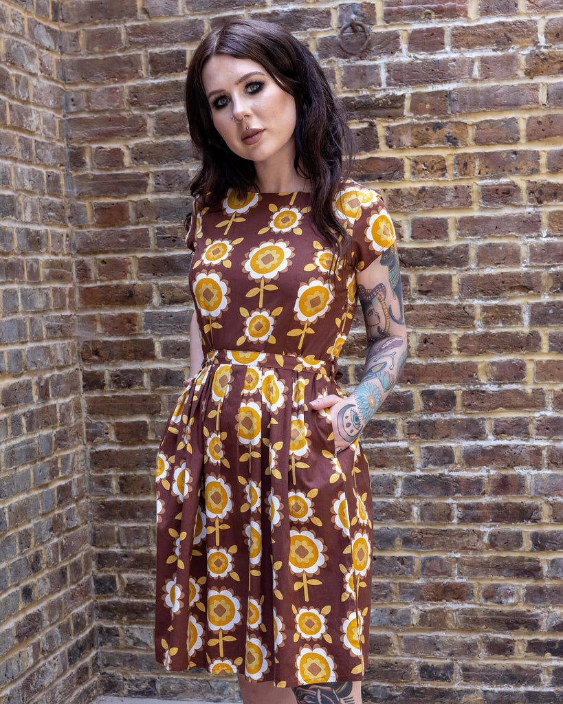 Florence a brown long haired white model with tattoos is wearing a brown retro tea dress with floral print against a brick wall.