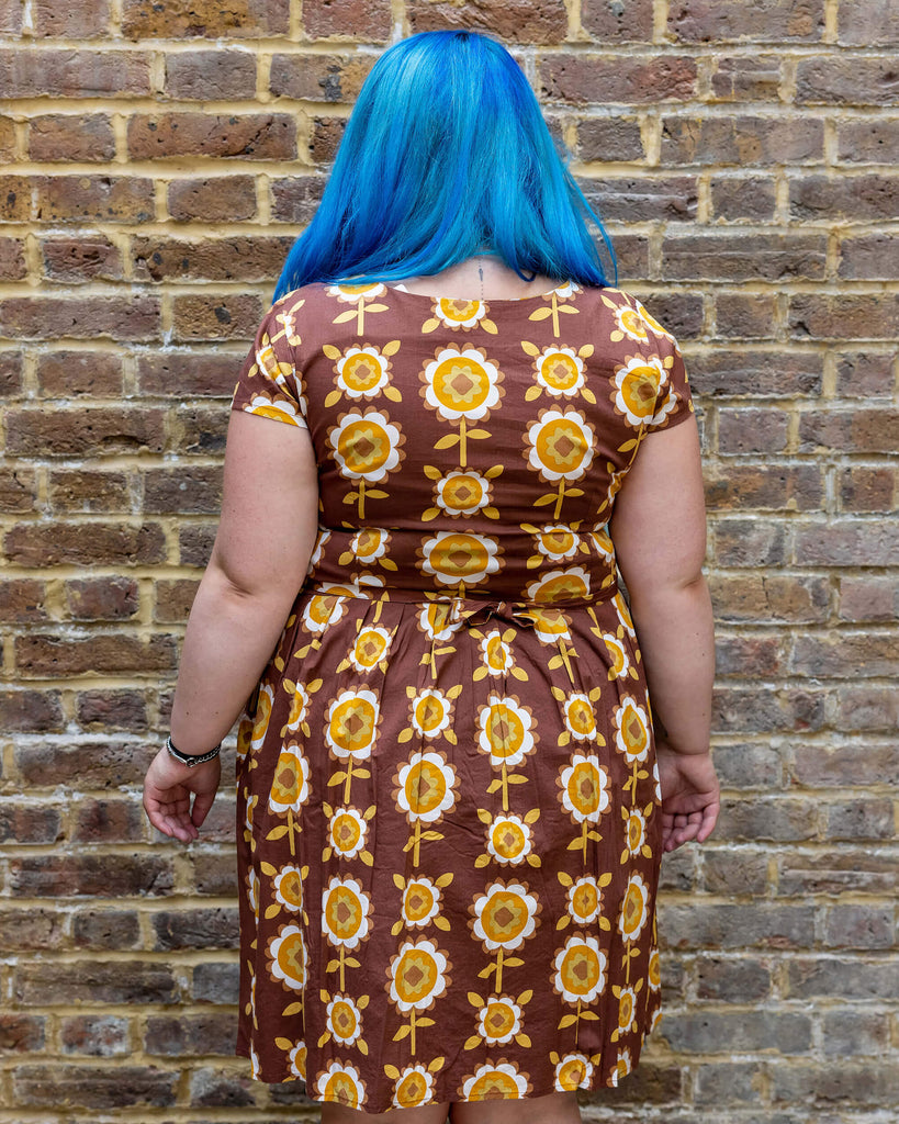 Georgina a blue long haired white plus sized model wearing a brown retro tea dress with floral print against a brick wall