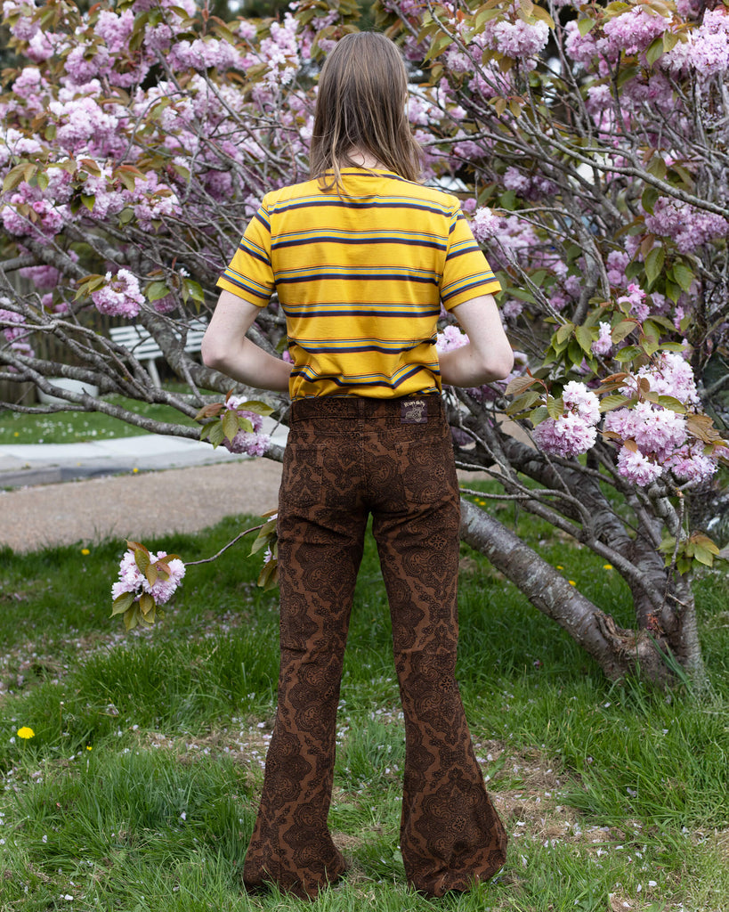 Jack, a male model with long blonde hair is stood outside in East Sussex by a blossom tree wearing Hendrix Tan Paisley Corduroy Retro Bell Bottom Flares paired with Gold Sunset Stripe Retro T Shirt. The trousers are a tan colour with an all over black paisley print. Jack is posing with his back to the camera with his arms down by his sides.