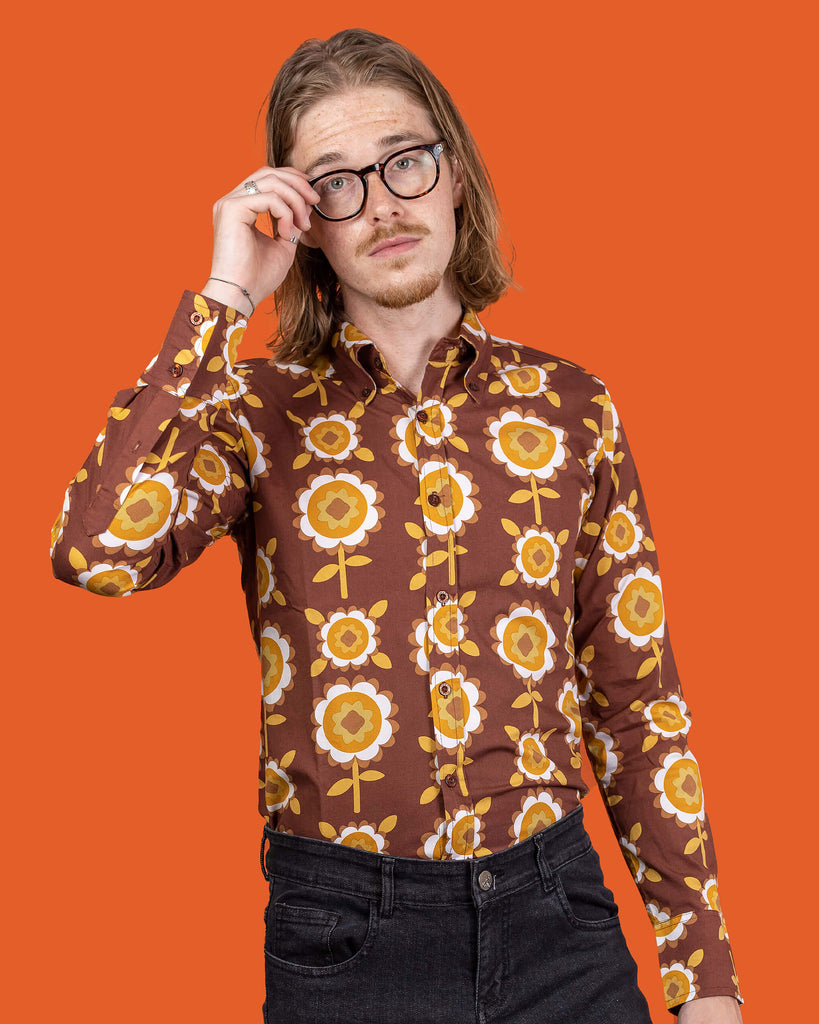 Jack a white male with shoulder length blonde hair and glasses in his 20's wearing a retro brown shirt with long selves and black jeans shot against an orange studio background.