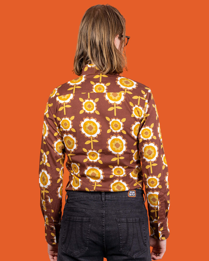 Jack a white male with shoulder length blonde hair and glasses in his 20's wearing a retro brown shirt with long selves and black jeans shot against an orange studio background.