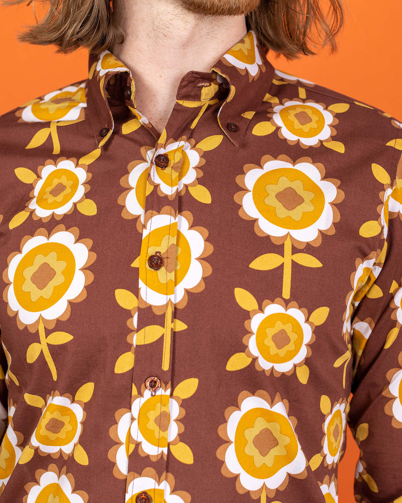 close up of Jack a white male with shoulder length blonde hair and glasses in his 20's wearing a retro brown shirt with long selves and black jeans shot against an orange studio background.