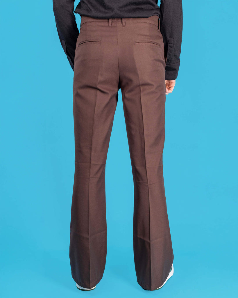 Jack is stood in front of a blue studio background wearing the Presley brown bell bottom trouser slacks with a long sleeve black tshirt and white trainers. They are facing away from the camera posing with both arms resting by their sides. Photo is cropped from the waist down.