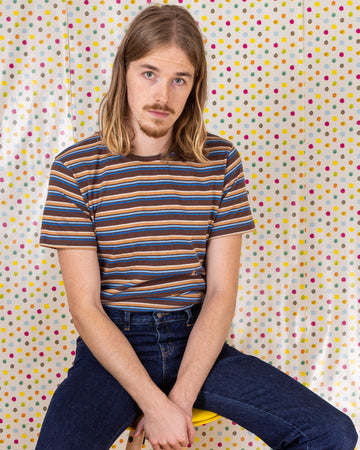 Model is sat in front of a multicoloured dot studio background wearing the brown stripe retro t-shirt with denim jeans. They are sat facing the camera with both hands resting by their sides and smiling.