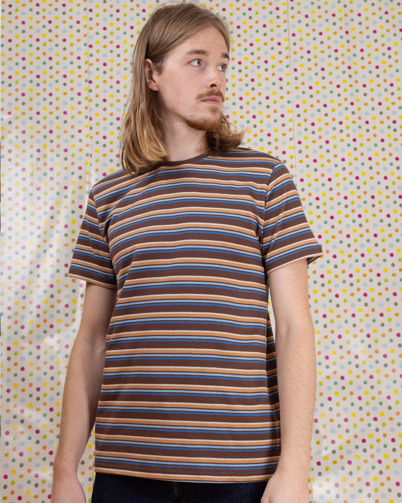Model is sat in front of a multicoloured dot studio background wearing the brown stripe retro t-shirt with denim jeans. They are sat facing the camera with both hands resting by their sides and smiling.