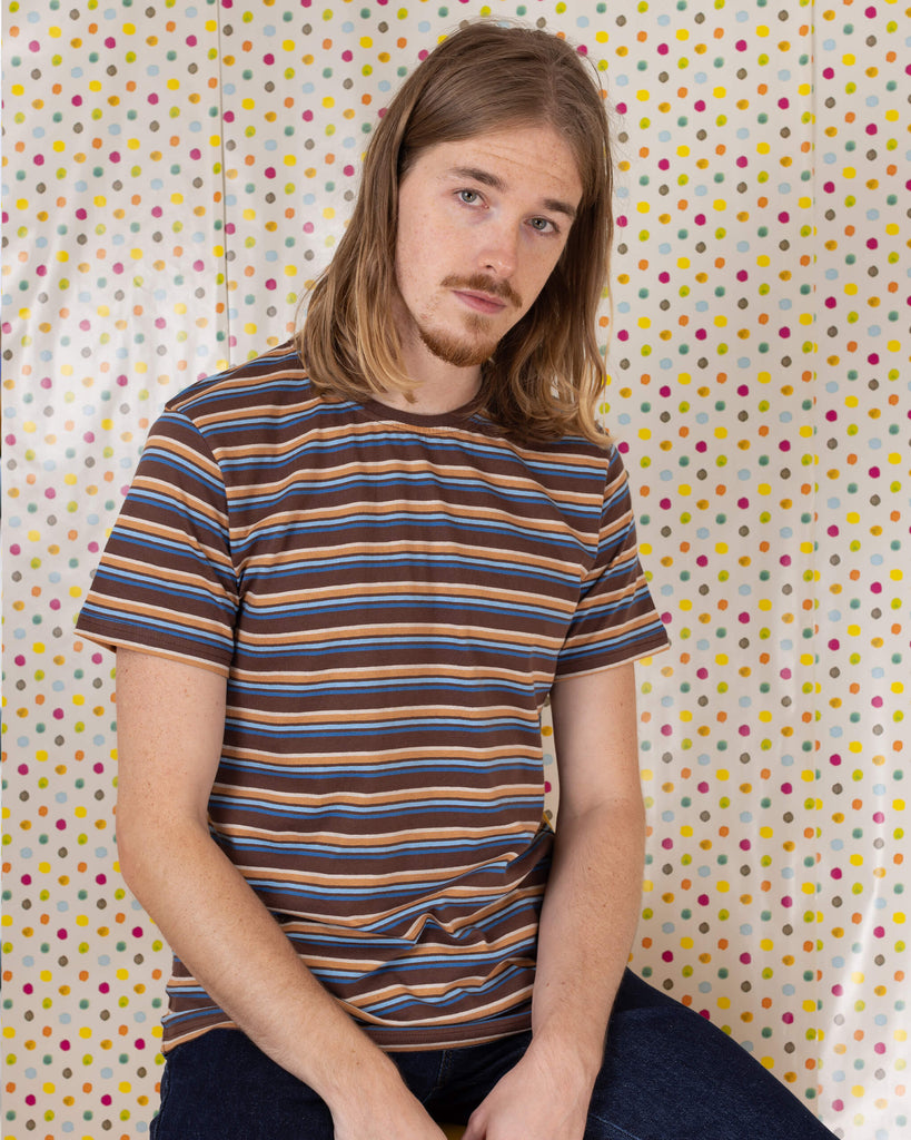 Model is sat in front of a multicoloured dot studio background wearing the brown stripe retro t-shirt with denim jeans. They are sat facing the camera with both hands resting by their sides and smiling.