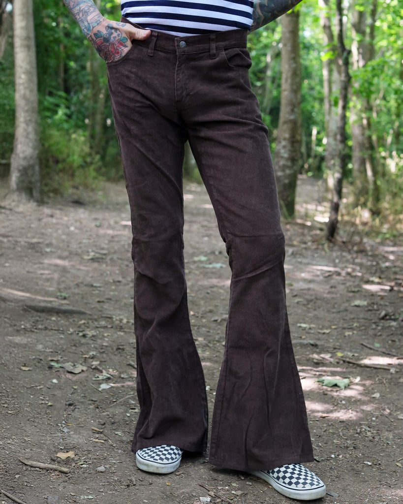 Model is stood in a forest area wearing the brown corduroy bell bottom super flares with a navy and white stripe t-shirt and checkered van shoes. They have one hand in their pocket with one hand behind their back, they are stood facing the camera with one leg slightly out to the side to show off the flares. Photo is cropped from the waist down.