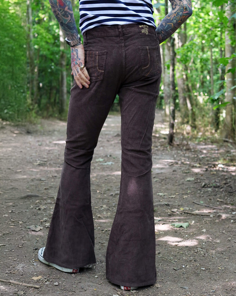 Model is stood in a forest area wearing the brown corduroy bell bottom super flares with a navy and white stripe t-shirt and checkered van shoes. They have one hand in their pocket with one hand behind their back, they are stood facing away from the camera with one leg slightly out to the side to show off the flares. Photo is cropped from the waist down.