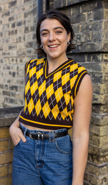 Freya a dark short haired white female model is wearing a brown and gold argyle knitted cropped tank with jeans stood in front of a brick wall
