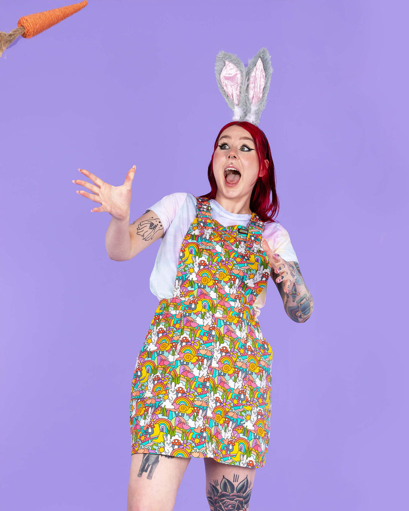 Florence a red haired tattooed femme white model wearing a Waffle Mama Design rainbow bunny print pinafore in multi colours with mushrooms, flowers, rainbows and clouds. She has paired this with a rainbow crop top, grey bunny ears and black trainers. She is facing forward posing on a purple background looking shocked as a carrot is thrown towards her.