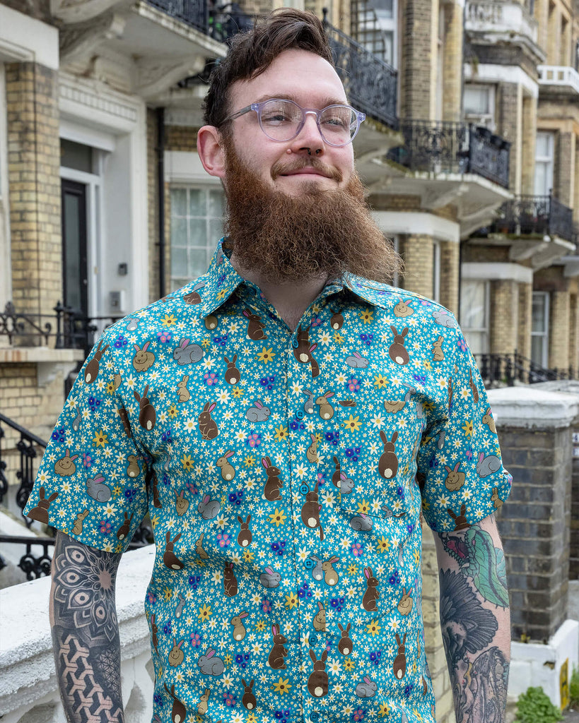 Dec is wearing the Bunny Meadow short sleeve blue shirt with floral pattern, worn with brown corduroy stretch flare trousers. Model is looking away from the camera outside of a street.