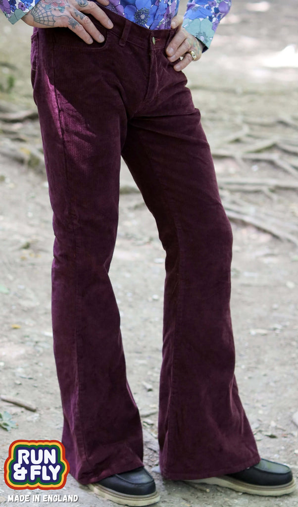 Model is stood outside in a forest area wearing the burgundy corduroy bell bottom super flares with a purple and blue floral shirt and dark brown boat shoes. They are facing slightly to the right and posing with both hands on their hips and one leg angled out to show off the flares. The photo is cropped from the waist down.