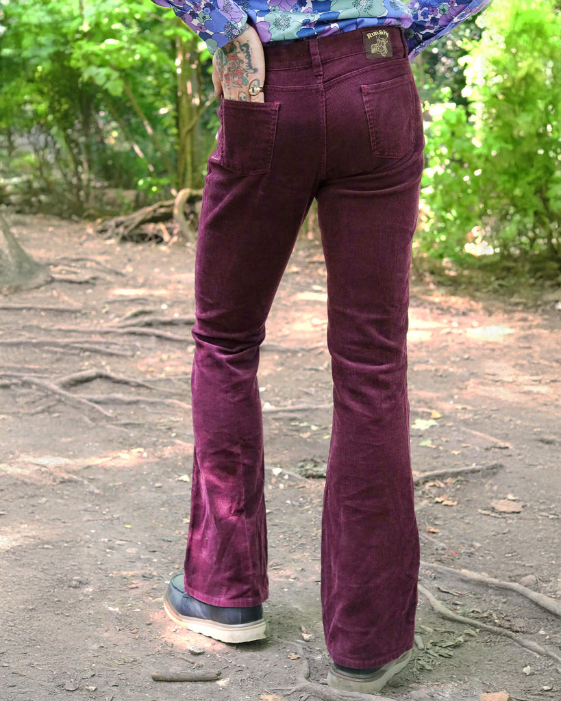 Model is stood in a forest area wearing the Burgundy boot cut flare cords with a purple floral shirt and brown boat shoes. The model is facing away from the camera posing with one leg out to the side to show off the flares. They have one hand resting by their side whilst the other is in their back pocket. Photo is cropped from the waist down.