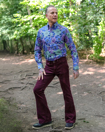 Model is stood in a forest area wearing the Burgundy boot cut flare cords with a purple floral shirt and brown boat shoes. The model is facing the camera posing with one leg out to the side to show off the flares with one hand on his thigh and the other behind his back. 