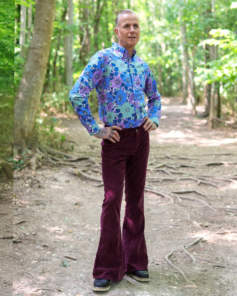 Model is stood outside in a forest area wearing the burgundy corduroy bell bottom super flares with a purple and blue floral shirt and dark brown boat shoes. They are facing slightly to the right and posing with both hands on their hips and one leg angled out to show off the flares. 