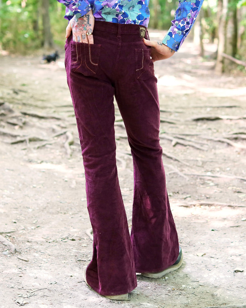 Model is stood outside in a forest area wearing the burgundy corduroy bell bottom super flares with a purple and blue floral shirt and dark brown boat shoes. They are facing away from the camera and posing with both hands on their hips and one leg angled out to show off the flares. The photo is cropped from the waist down.