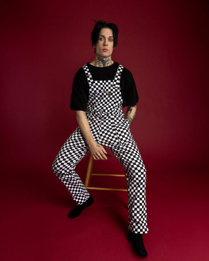 The Black & White Checkerboard Stretch Twill Dungarees on a gothic style male model with tattoos and piercings. He has black hair with purple eyeshadow and is sat on a wooden stool on a red background, he has one hand in the pocket and the other hand resting between his legs, he is smising to camera. The slow fashion dungarees are black and white check print with retro rollers, matching turn ups and five different pockets.