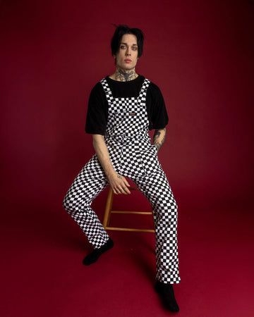 The Black & White Checkerboard Stretch Twill Dungarees on a gothic style male model with tattoos and piercings. He has black hair with purple eyeshadow and is sat on a wooden stool on a red background, he has one hand in the pocket and the other hand resting between his legs, he is smising to camera. The slow fashion dungarees are black and white check print with retro rollers, matching turn ups and five different pockets.