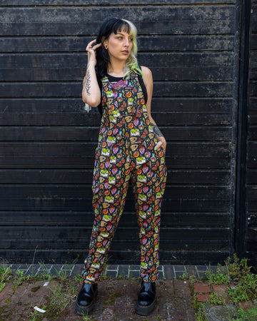 Person posing in Run & Fly dungarees with hands in pockets, featuring a colourful print of magical elements. The person has black and blonde hair, multiple piercings, and a tattoo on their arm, adding to the edgy, alternative look.