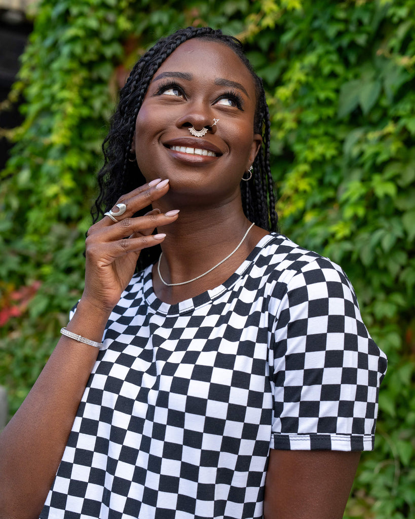 Close-up of a model smiling while wearing a black and white checkered dress from Run & Fly. The model has long braided hair, silver jewellery, and a crescent moon nose ring, with a backdrop of green leaves. The playful expression and stylish outfit reflect the ethical, sustainable values of the slow fashion brand.