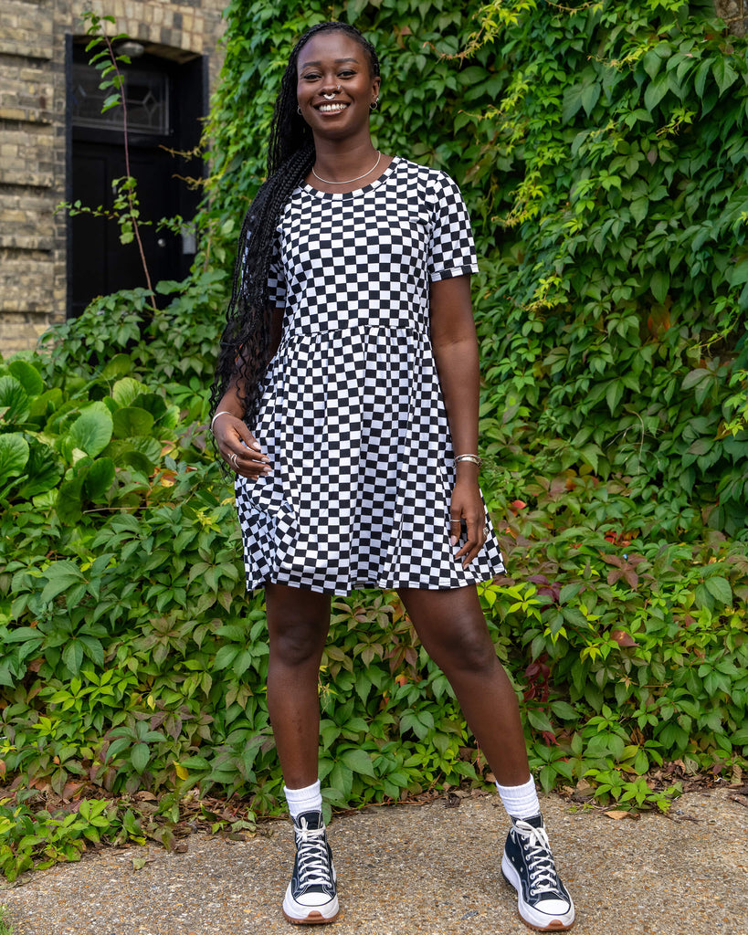 Model wearing a black and white checkered dress by Run & Fly, smiling while standing in front of lush green plants. The relaxed-fit dress with short sleeves highlights the brand’s commitment to ethical and sustainable slow fashion. Black platform trainers complete the look.