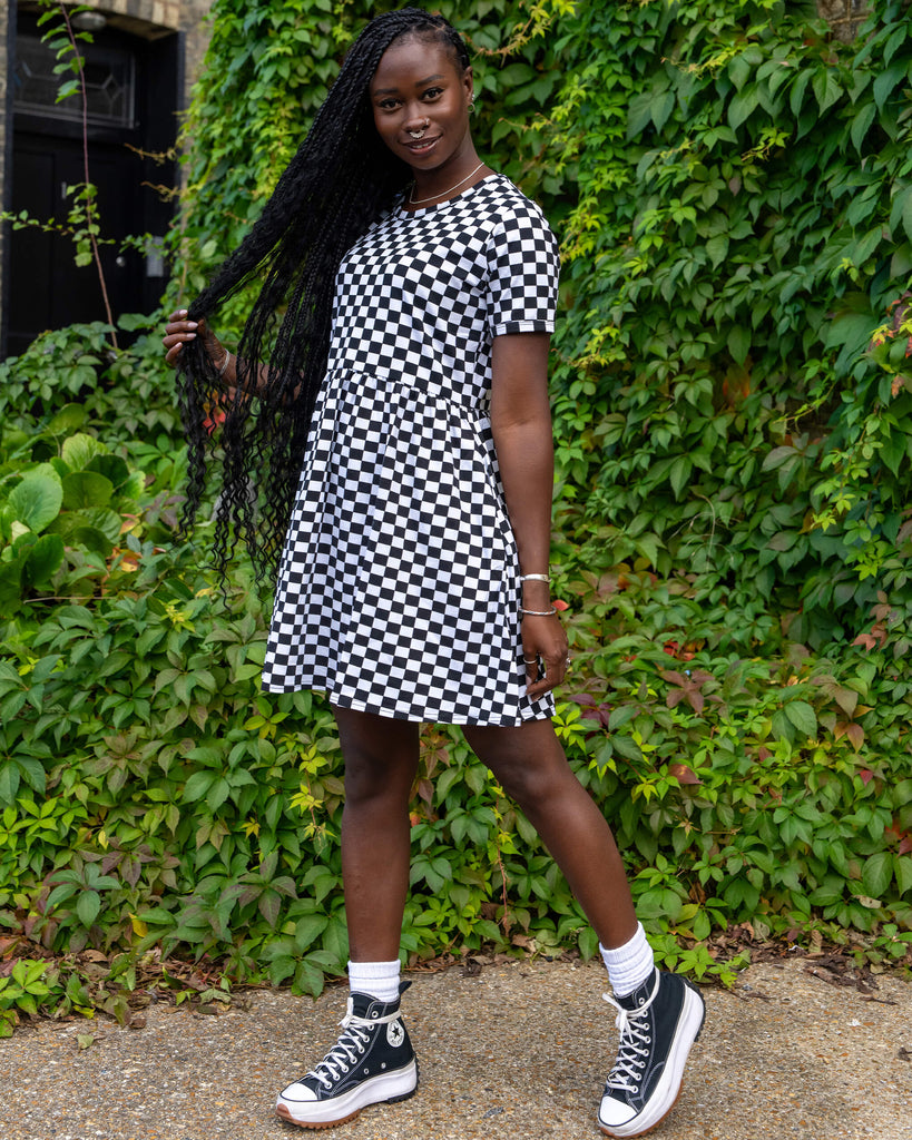 Run & Fly's black and white checkered dress is displayed by a model pulling their long braided hair to the side. The dress has a relaxed fit and short sleeves, styled with platform trainers. The natural setting reflects the sustainable and ethical values of the brand.
