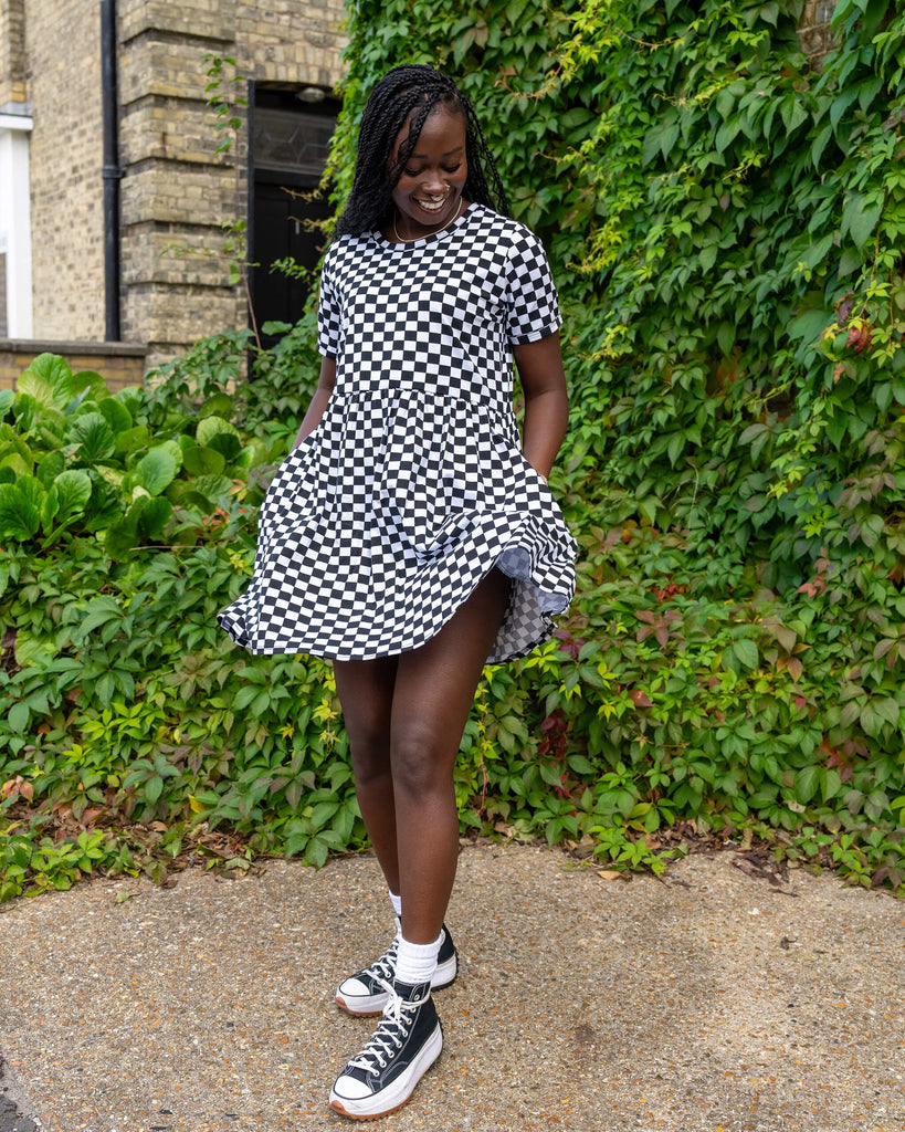 The model in a black and white checkered dress from Run & Fly is smiling and twirling. The loose dress features a flattering fit and short sleeves, paired with black trainers and white socks. Surrounded by greenery, this highlights the brand’s ethical and slow fashion ethos.