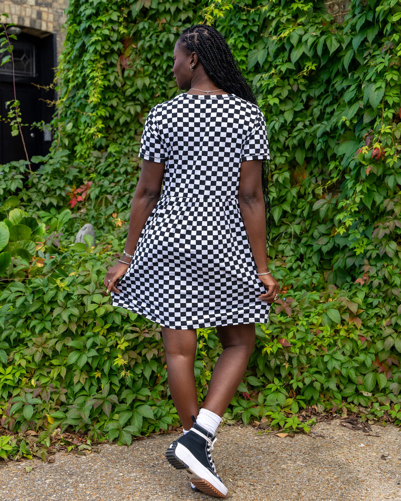 Model in a black and white checkered dress from Run & Fly, standing with their back facing the camera. The relaxed-fit dress falls above the knee, and the model is wearing black platform trainers. Shot in front of greenery, this showcases the eco-friendly, ethical slow fashion focus of the brand.