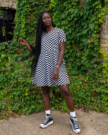 The model wearing a black and white checkered dress from Run & Fly strikes a fun pose, holding their long braided hair. The knee-length dress and black trainers pair well with the natural backdrop, reflecting the slow fashion brand's sustainable focus