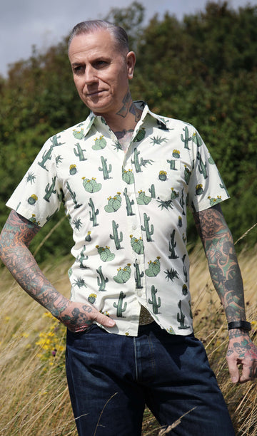 A heavily tattooed model is stood outside in a tall grass field wearing the cactus short sleeve shirt with denim jeans. He is facing towards the camera with one hand in his front pocket with the other resting by his side, he is looking off to the left.