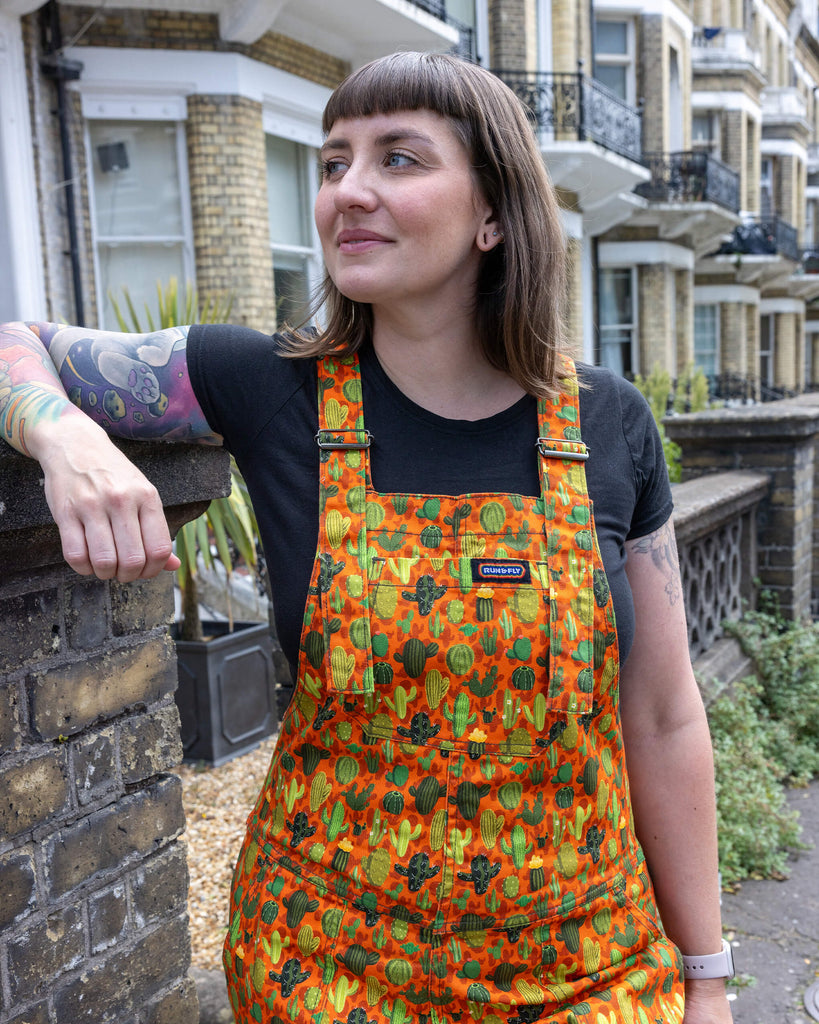 Laura a white femme tattooed model is wearing orange dungarees with all over cactus print paired with a black t-shirt while smiling looking off to one side leant against a wall outdoors in Hove.