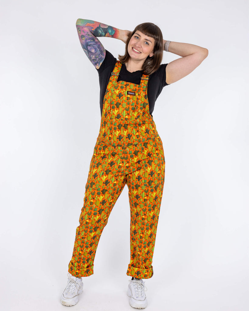 Laura a white femme tattooed model is wearing orange dungarees with all over cactus print paired with a black t-shirt and white trainers. Laura is smiling at the camera with her hands behind her head in front of a white background.