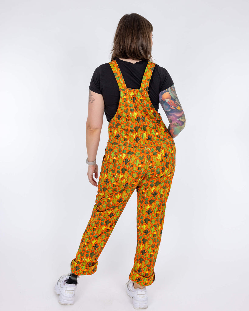 Laura a white femme tattooed model is wearing orange dungarees with all over cactus print paired with a black t-shirt and white trainers. Laura is stood with her back to the camera in front of a white background.