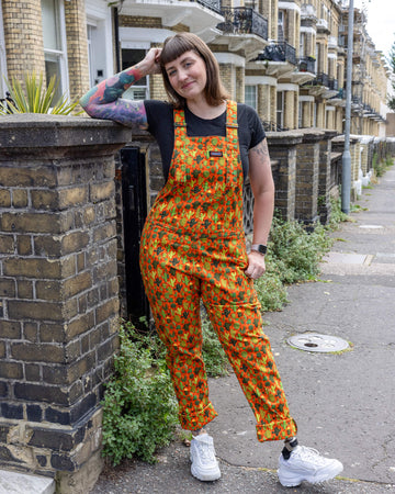 Laura a white femme tattooed model is wearing orange dungarees with all over cactus print paired with a black t-shirt and white trainers. Laura is leaning against a wall with one leg turned outwards outdoors in Hove.