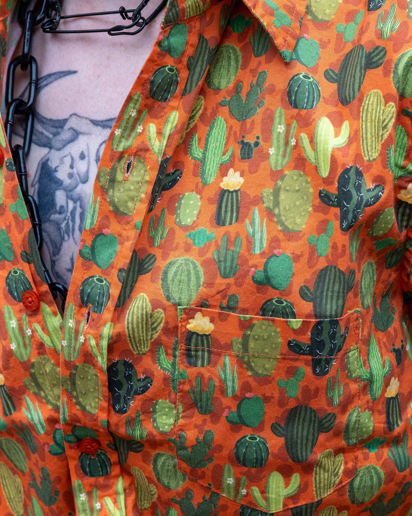 Close up of Jamie a trans masc white man with blonde short hair and black tattoos is wearing a cactus print orange based shirt and black denim shorts. 