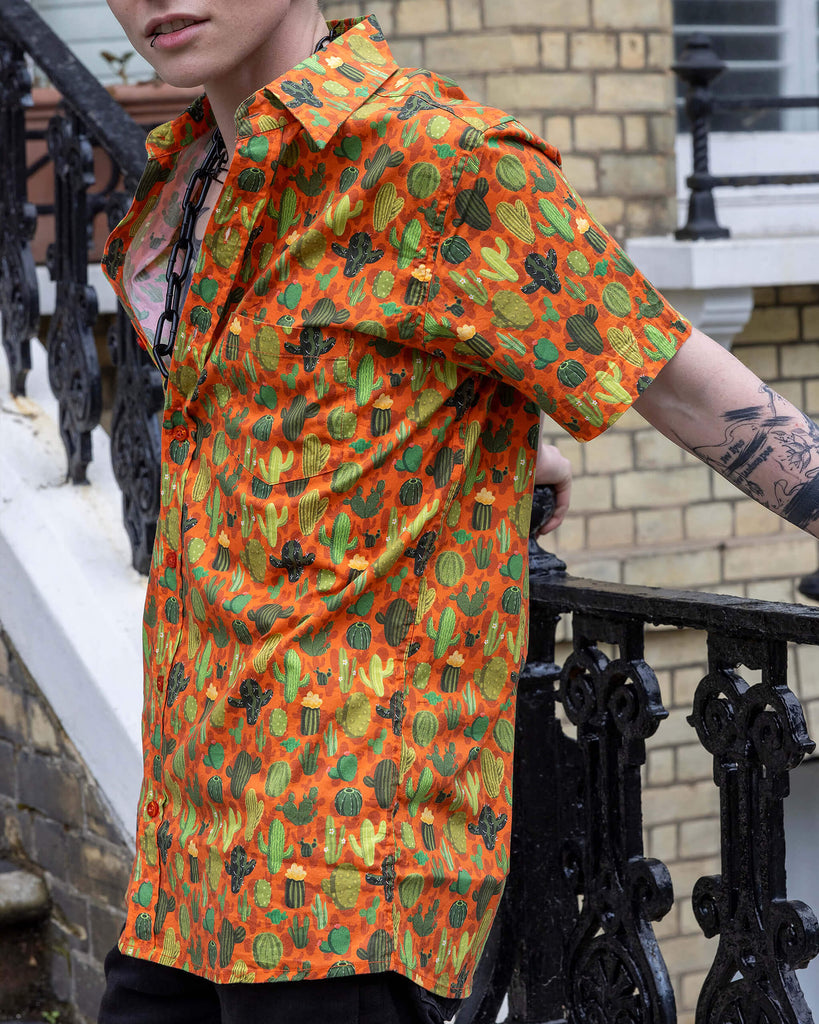 Jamie a trans masc white man with blonde short hair and black tattoos is wearing a cactus print orange based shirt and black denim shorts. He is stood on a set of steps in Hove.