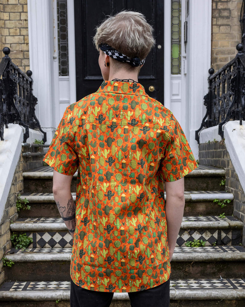 Back view of Jamie a trans masc white man with blonde short hair and black tattoos is wearing a cactus print orange based shirt and black denim shorts. He is stood facing away on a set of steps in Hove.