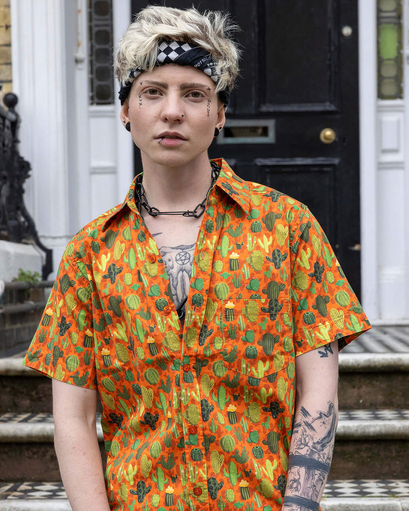 Jamie a trans masc white man with blonde short hair and black tattoos is wearing a cactus print orange based shirt and black denim shorts. He is stood on a step in Hove facing forwards holding the bottom of the shirt.