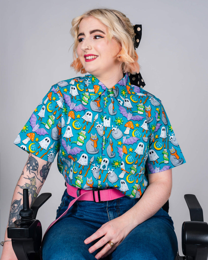 A smiling person with blonde and pink hair wearing a vibrant blue shirt with Halloween motifs such as ghosts and mushrooms, designed by ethical fashion brand Run & Fly.