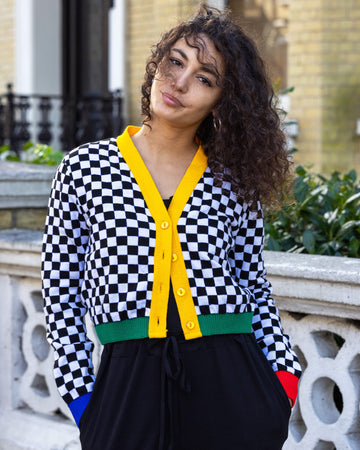 The Colour Block Checkerboard Cropped Cardigan worn by a femme model with curly brown hair with black trousers. She is facing forward smiling slightly to camera with both hands in her pockets. The v neck crop cardigan with four yellow circle buttons is a black and white checkerboard print with a yellow centre trim, a green hem trim, blue right sleeve hem and red left sleeve hem.