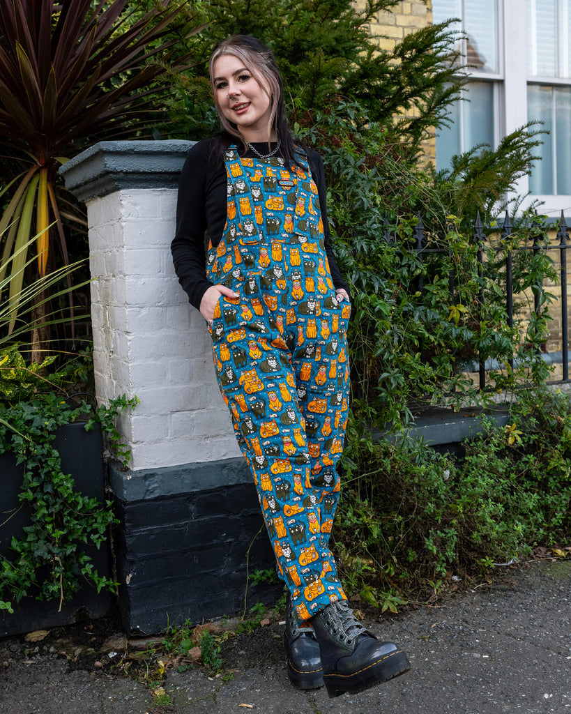 Model leaning against a white brick pillar, wearing teal dungarees with a playful orange and black cat design by Waffle Mama Design, looking relaxed and casual.